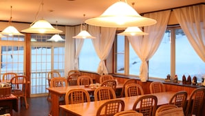A dining area with wooden tables and chairs, large windows with curtains, and hanging pendant lights. - Pension Tama House (Yuzawa)