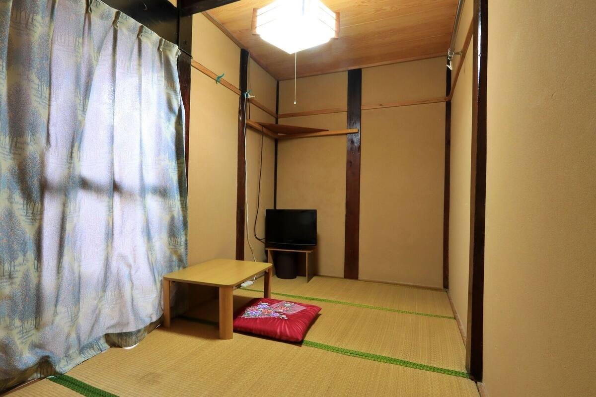 Une pièce avec un sol recouvert de tatamis, une petite table en bois, un coussin rouge et un téléviseur posé sur un meuble.