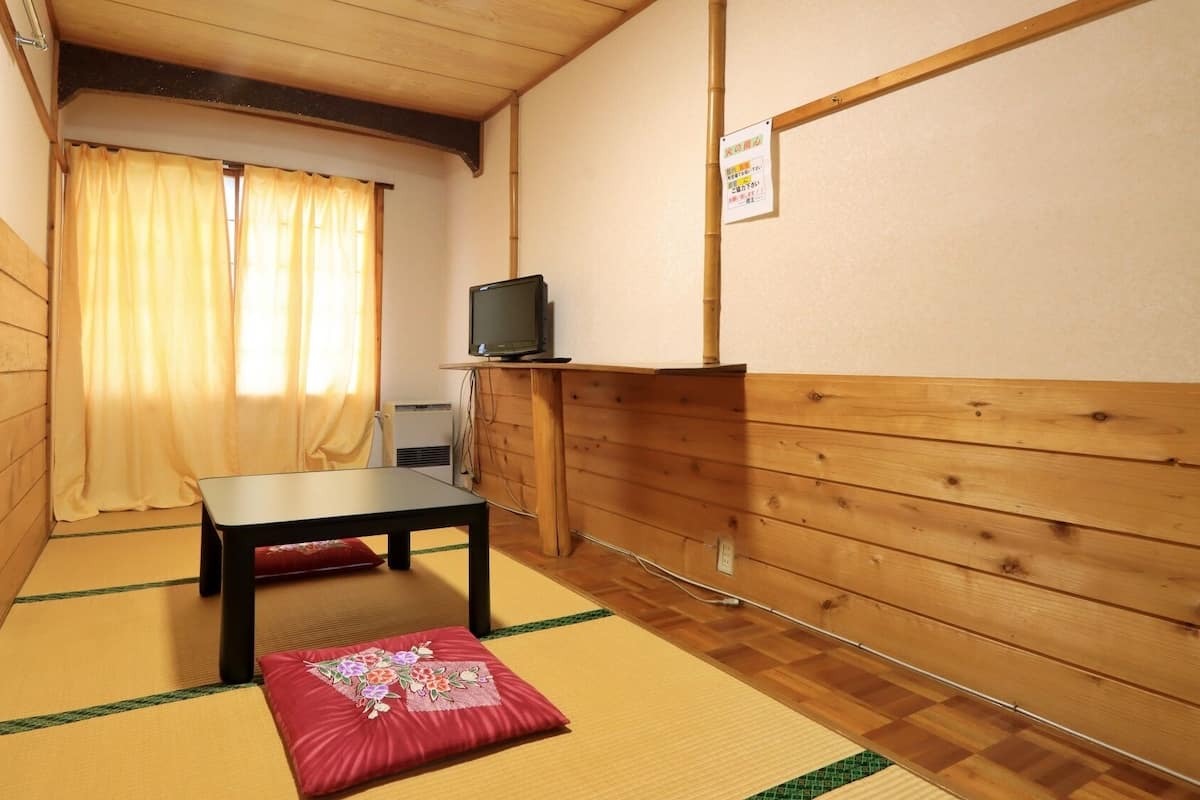 Une pièce de style japonais traditionnel, avec un sol recouvert de tatamis, une table basse, un téléviseur posé sur un meuble en bois et une porte coulissante avec des rideaux.
