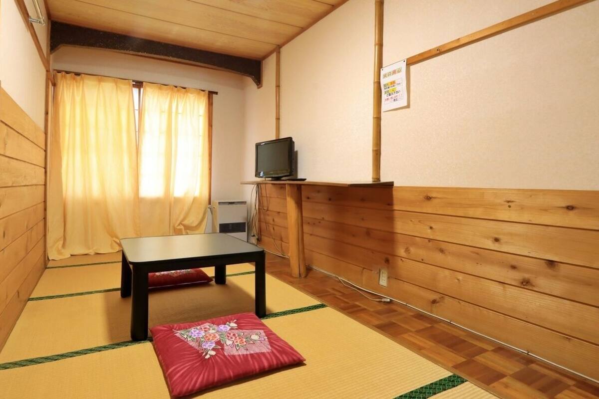 Une pièce étroite aux murs en bois, dotée d’un sol recouvert de tatamis, d’une petite table, d’un téléviseur et d’un coussin rouge.