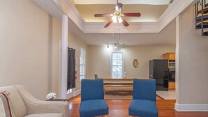 Un salon avec un canapé beige, deux fauteuils bleus, une table basse en bois, un ventilateur de plafond, et une cuisine en arrière-plan.