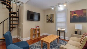 Un salon avec un escalier en bois, un téléviseur à écran plat, un canapé, une table basse, une lampe et un tableau accroché au mur.
