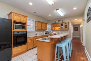 A kitchen with wooden cabinets, a refrigerator, a microwave, an oven, a sink, and a bar stool area. - Downtown, Moments from Riverwalk-Broadway Bungalow (Columbus)