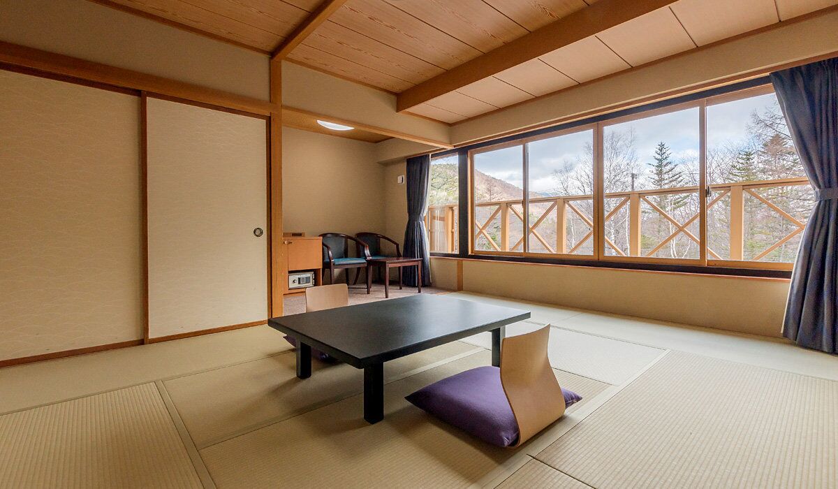 Une pièce de style japonais traditionnel, avec un sol recouvert de tatamis, une table basse, des chaises et une grande fenêtre donnant sur la forêt.