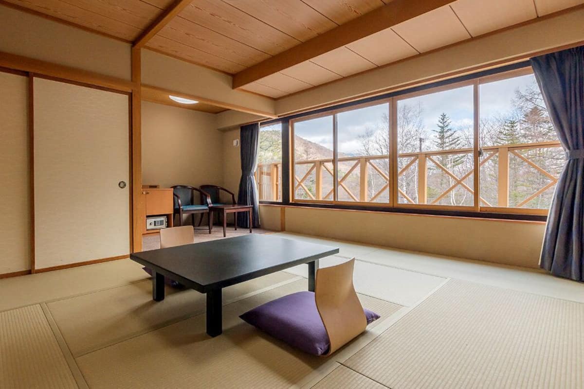Ein Raum mit Tatami-Matten, einem niedrigen Tisch, Stühlen und Blick durch ein großes Fenster.