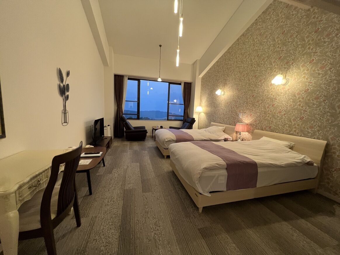 A hotel room with a bed, desk, chair, and a view through the window.