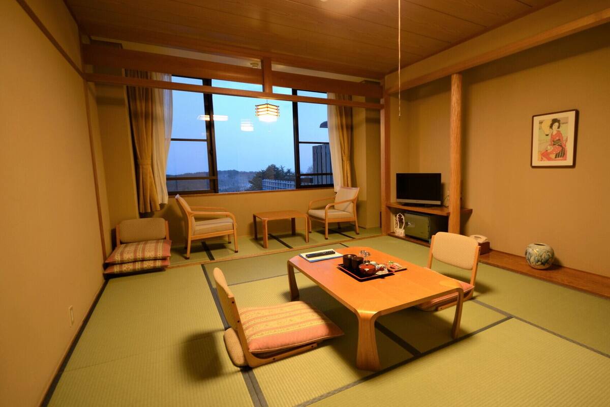 A traditional Japanese-style room with tatami flooring, a low table, two chairs, a television, and a view through large windows.