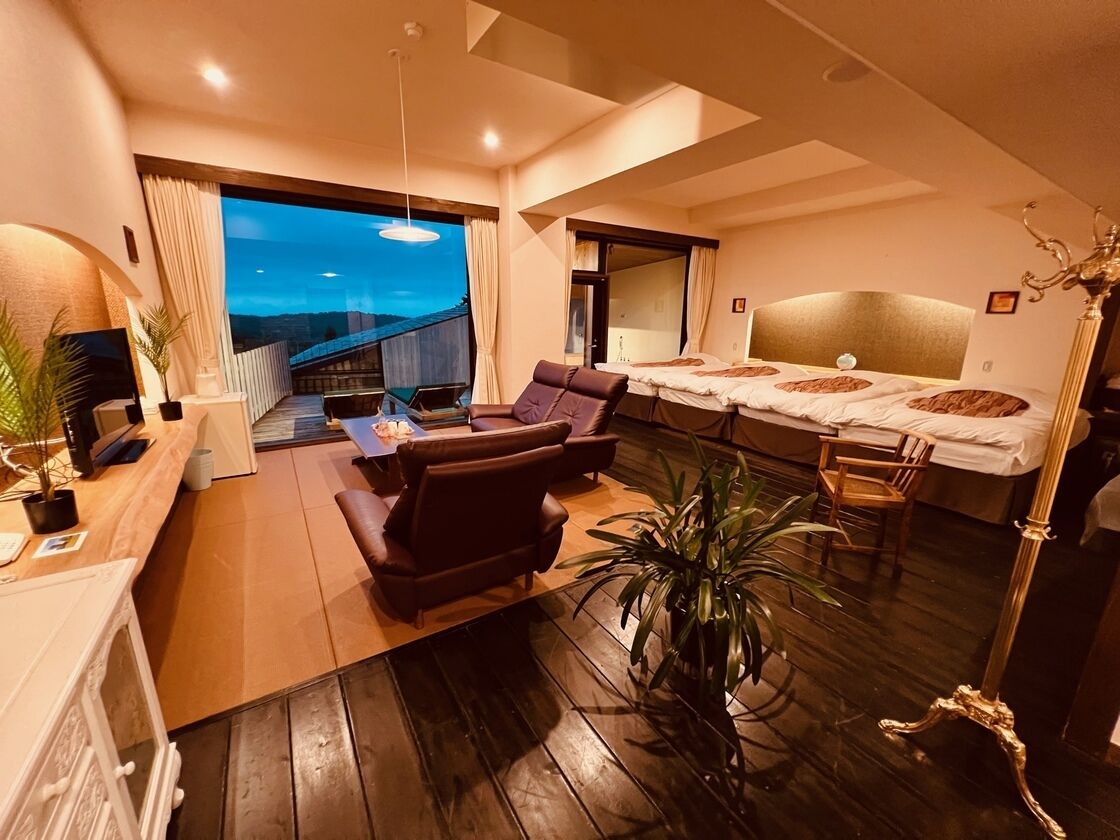 Habitación de hotel moderna con una cama grande, zona de estar con sofá y sillones, mesa de comedor y balcón con vistas.