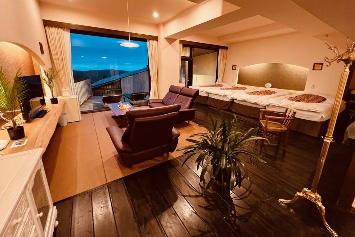 A modern hotel room with a large bed, a seating area with a sofa and chairs, a dining table, and a balcony with a view.