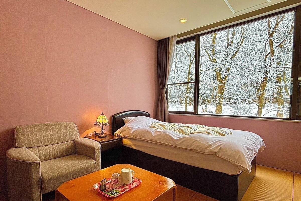 A hotel room with a bed, a chair, a nightstand with a lamp, and a window with a view of snow-covered trees.