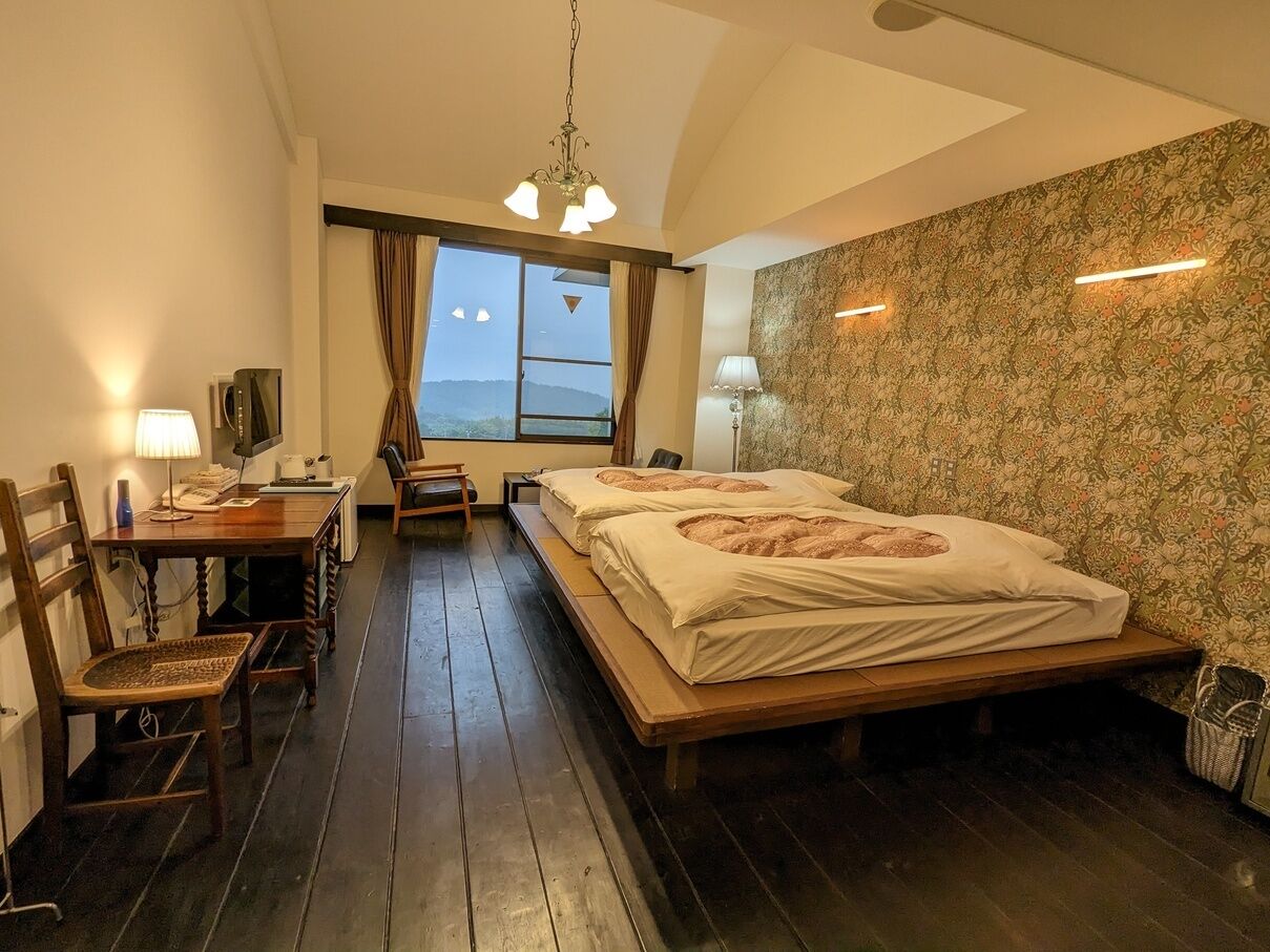 A bedroom with a bed, a desk, a chair, a window with curtains, and a view of the outdoors.