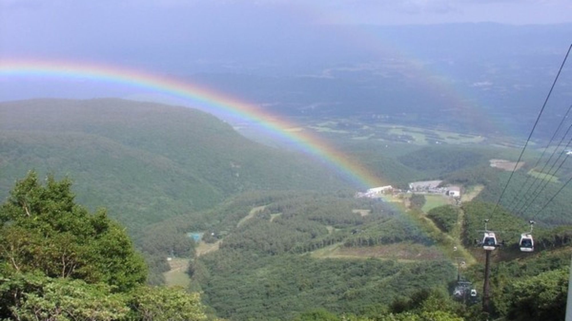 Eine Berglandschaft mit Regenbogen, einem Tal und einer kleinen Siedlung mit Gebäuden.