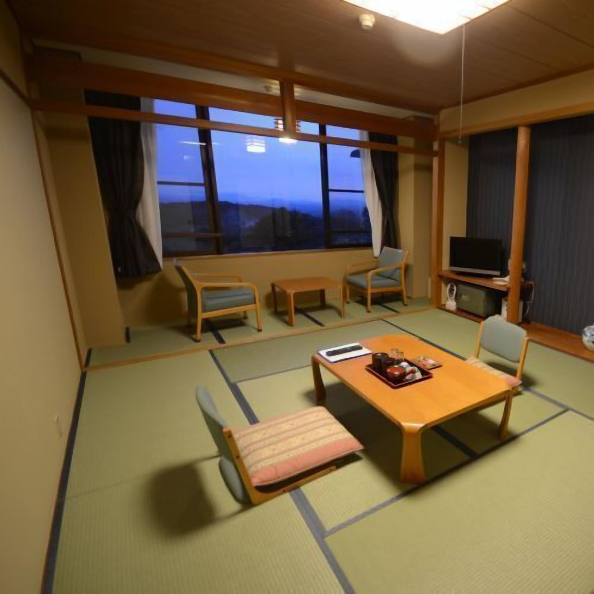 A traditional Japanese room with tatami flooring, a low table with a cushion, two chairs, a television, and a view of the mountains.