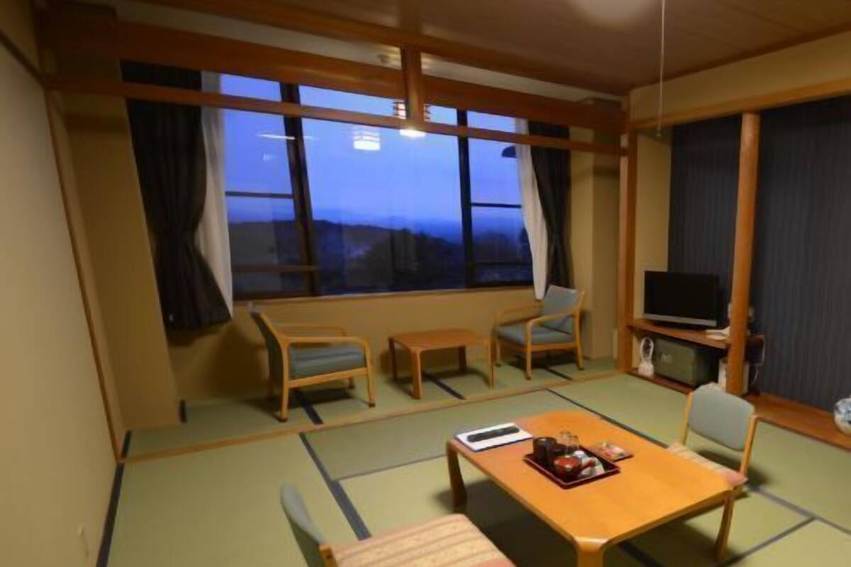 A traditional Japanese room with tatami flooring, a low table with a cushion, two chairs, a television, and a view of the mountains.