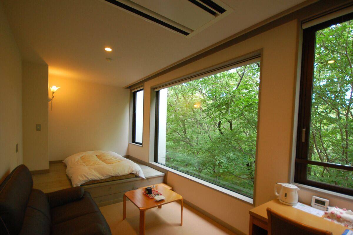 A room with a bed, a sofa, a coffee table, a desk, and a window with a view of trees.