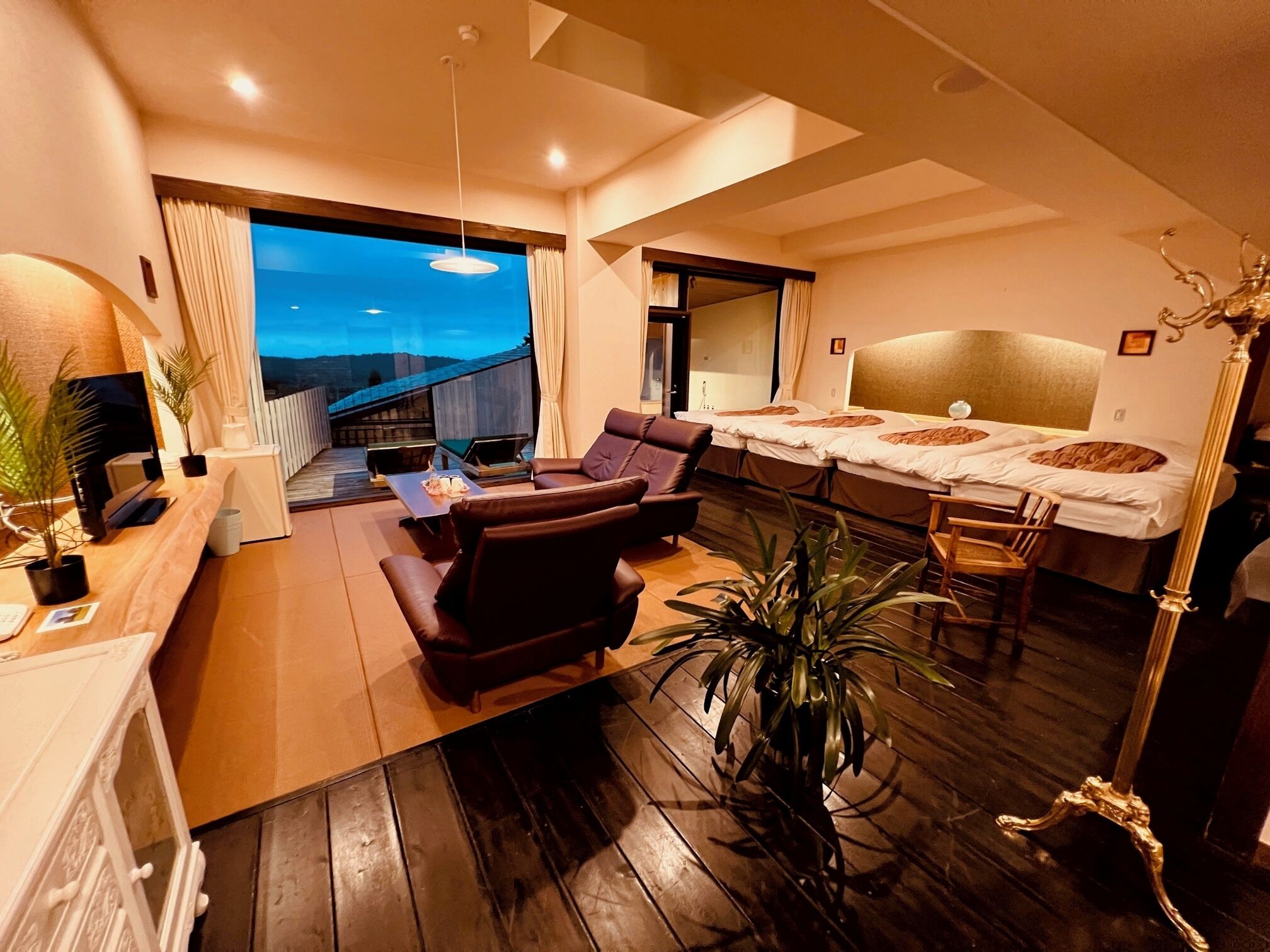 Una habitación de hotel moderna con piso de madera, un sofá, un escritorio con una computadora, una cama con cabecera y vista a un edificio a través de un ventanal grande.