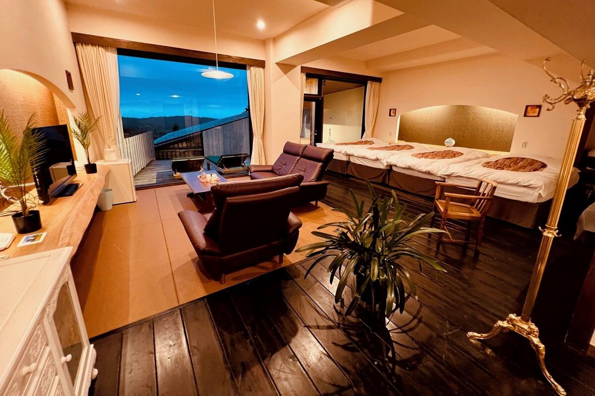 A modern hotel room with a wooden floor, a sofa, a desk with a computer, a bed with a headboard, and a view of a building through a large window.