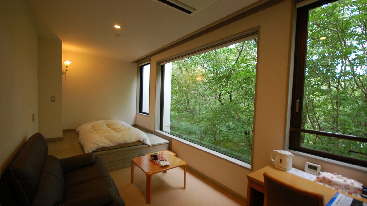 A room with a bed, a sofa, a small table, a window with a view of trees, and a wall-mounted light.