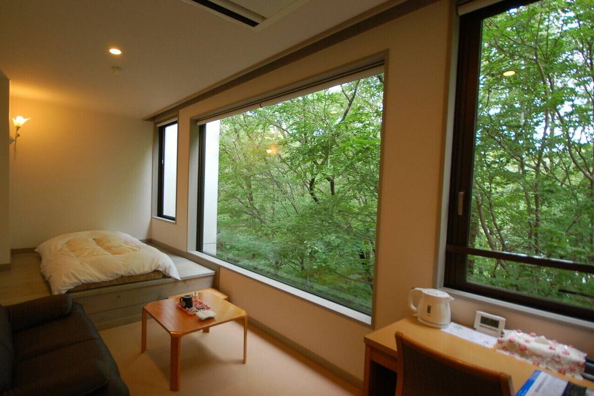 A room with a bed, a sofa, a small table, a window with a view of trees, and a wall-mounted light.