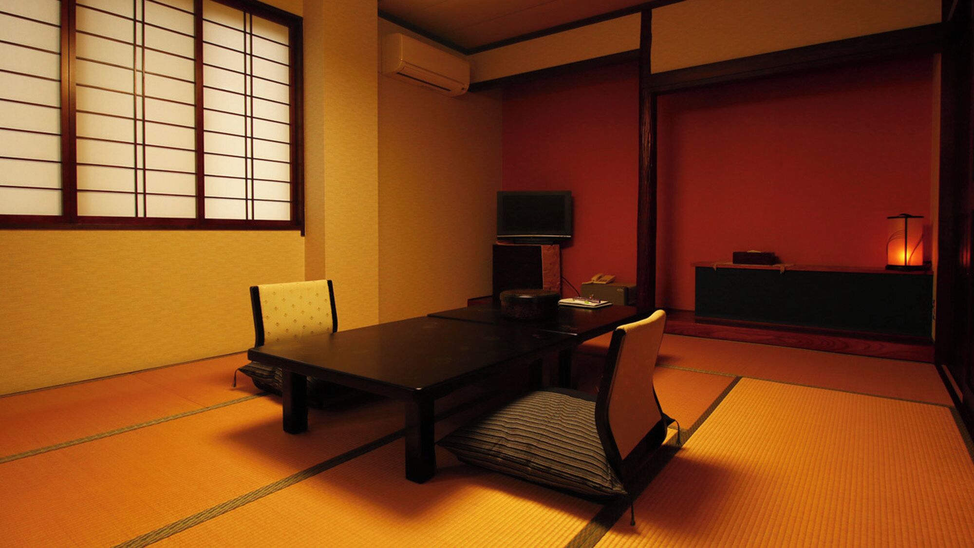 Una habitación tradicional japonesa con suelo de tatami, una mesa baja, sillas, un televisor y una pared roja.
