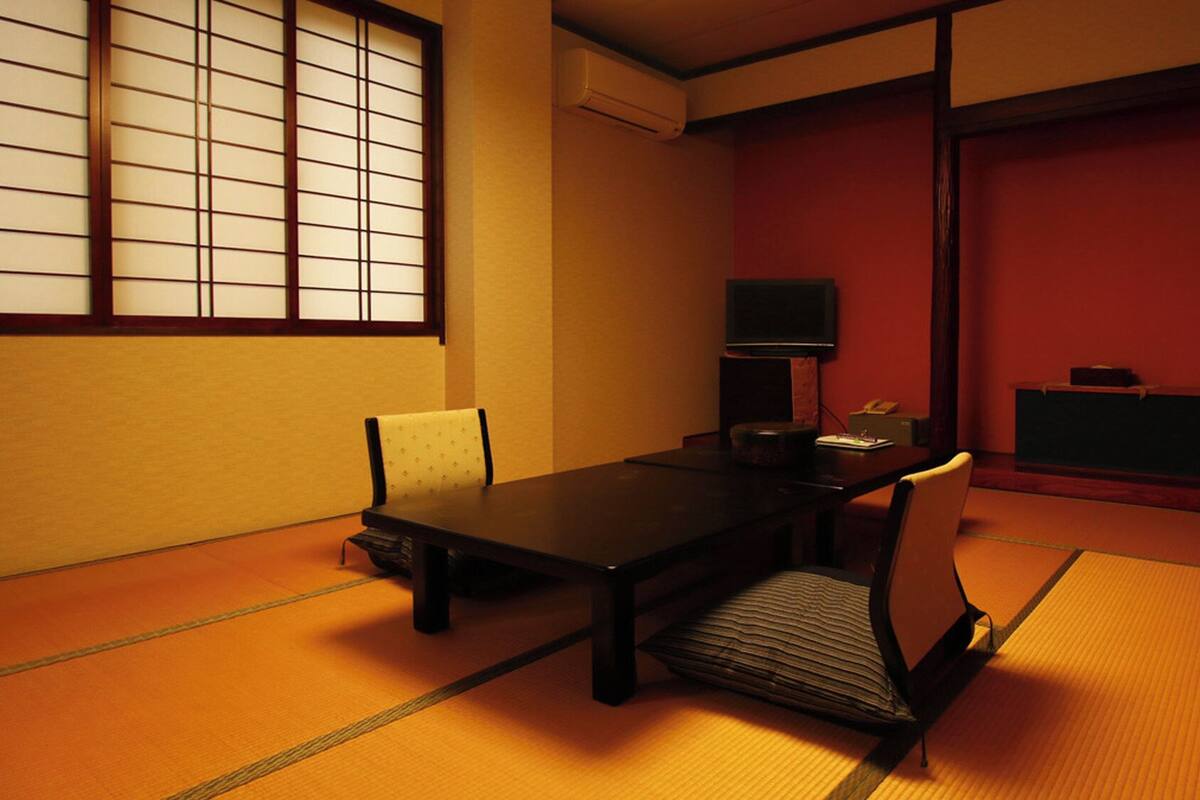 Une pièce de style japonais traditionnel, avec un sol recouvert de tatamis, une table basse, des chaises, un téléviseur et un mur rouge.