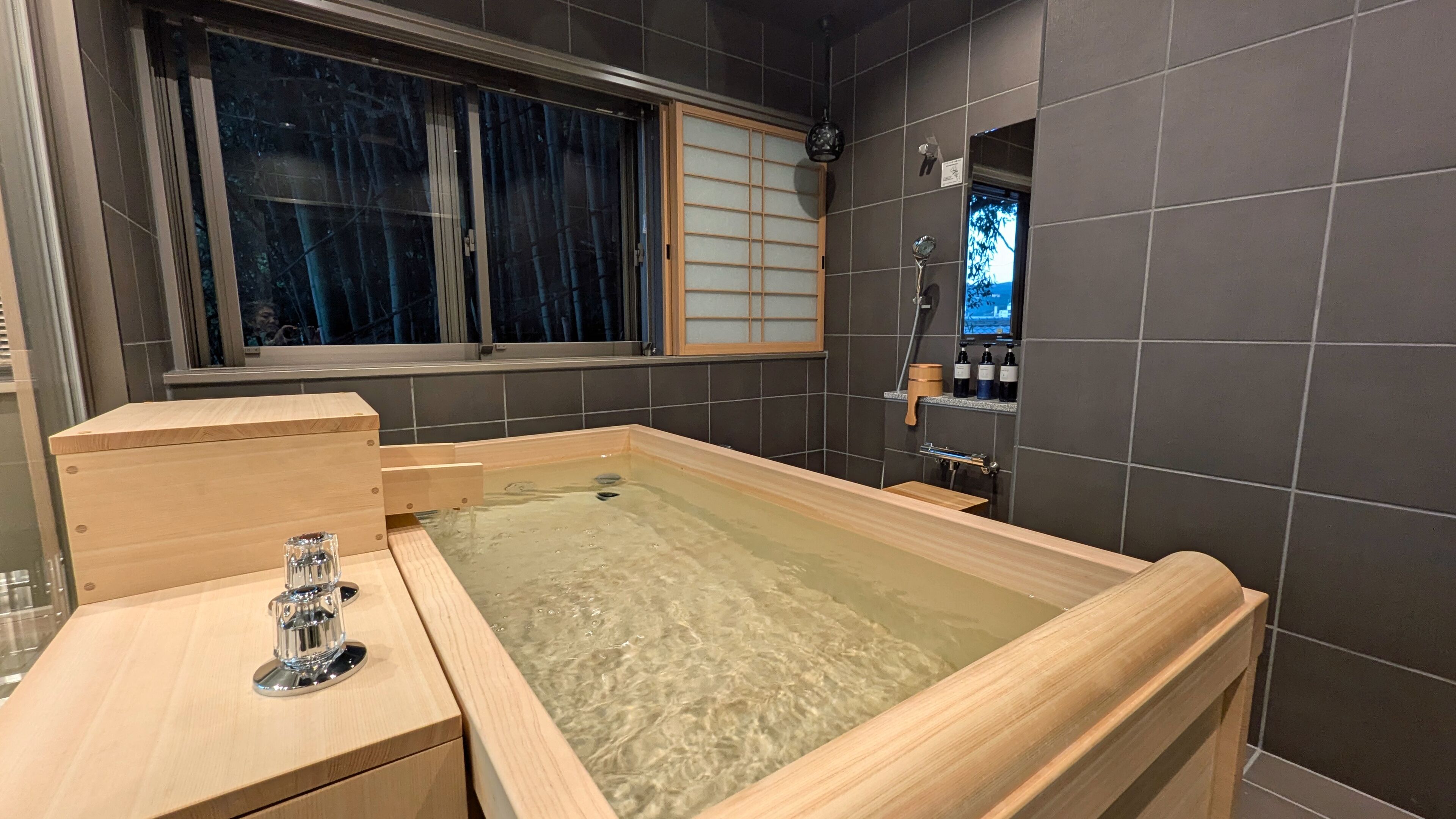 A modern bathroom with a wooden bathtub, a glass shower enclosure, and a window with a view.