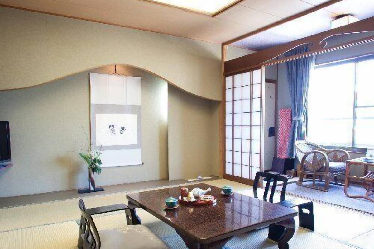 A traditional Japanese room with tatami flooring, a low table with cushions, a TV, and a sliding door leading to a balcony with wicker furniture.