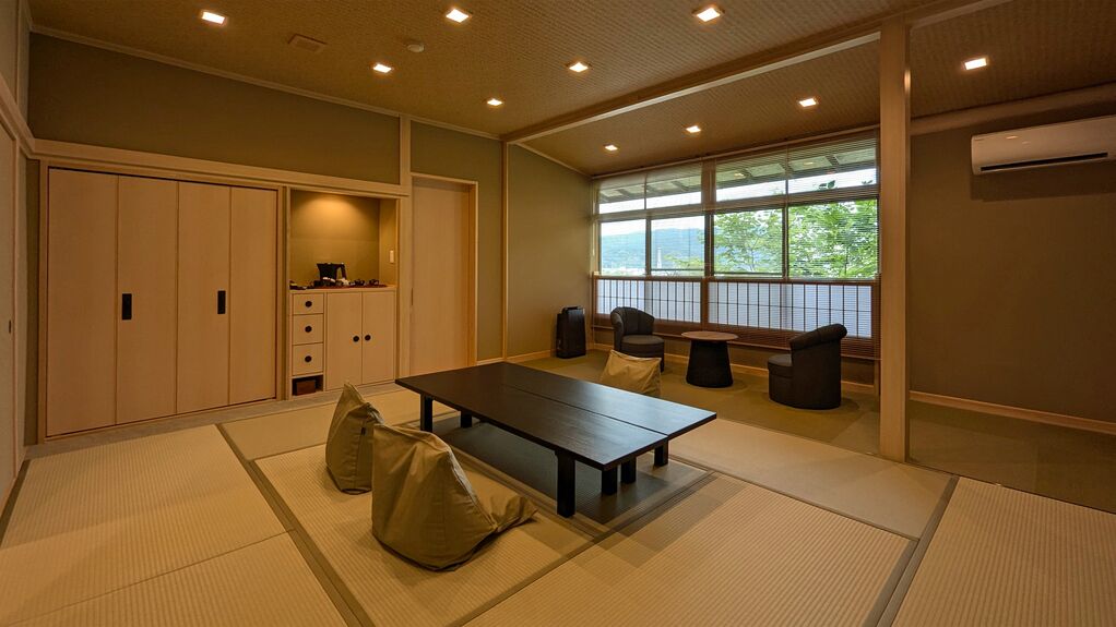Ein Zimmer mit Tatami-Matten, einem niedrigen Tisch, Stühlen, einem Kleiderschrank und einem Fenster mit Aussicht.
