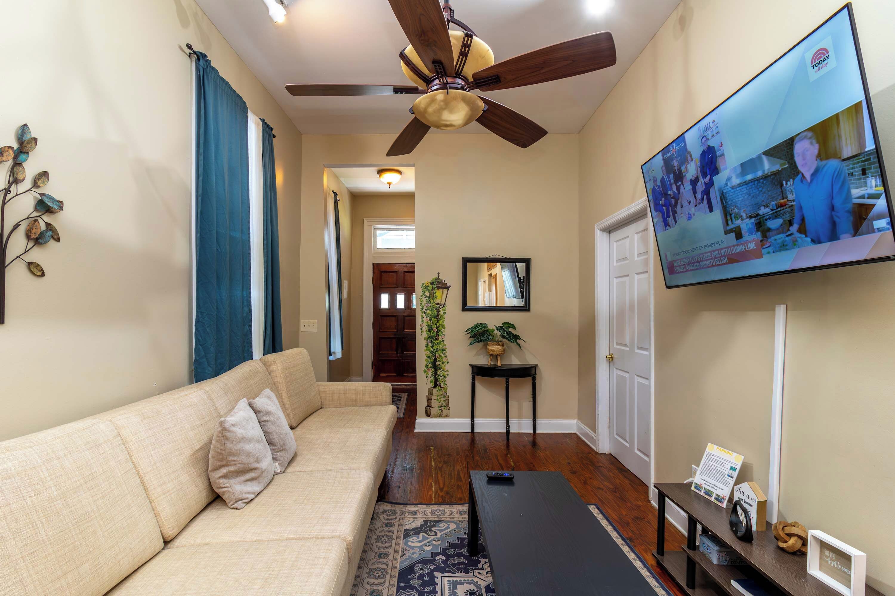 A living room with a sofa, a coffee table, a TV mounted on the wall, a ceiling fan, and a staircase leading to another room.