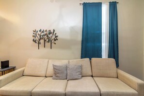 A beige sofa with two cushions, a wall-mounted decorative leaf art piece, and a window with blue curtains. - Closest Historic BnB to Ft Moore-Americana Cottage (Columbus)