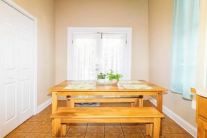 Une table de salle à manger en bois, accompagnée d’un banc, d’une plante en pot et d’une fenêtre ornée de rideaux légers.