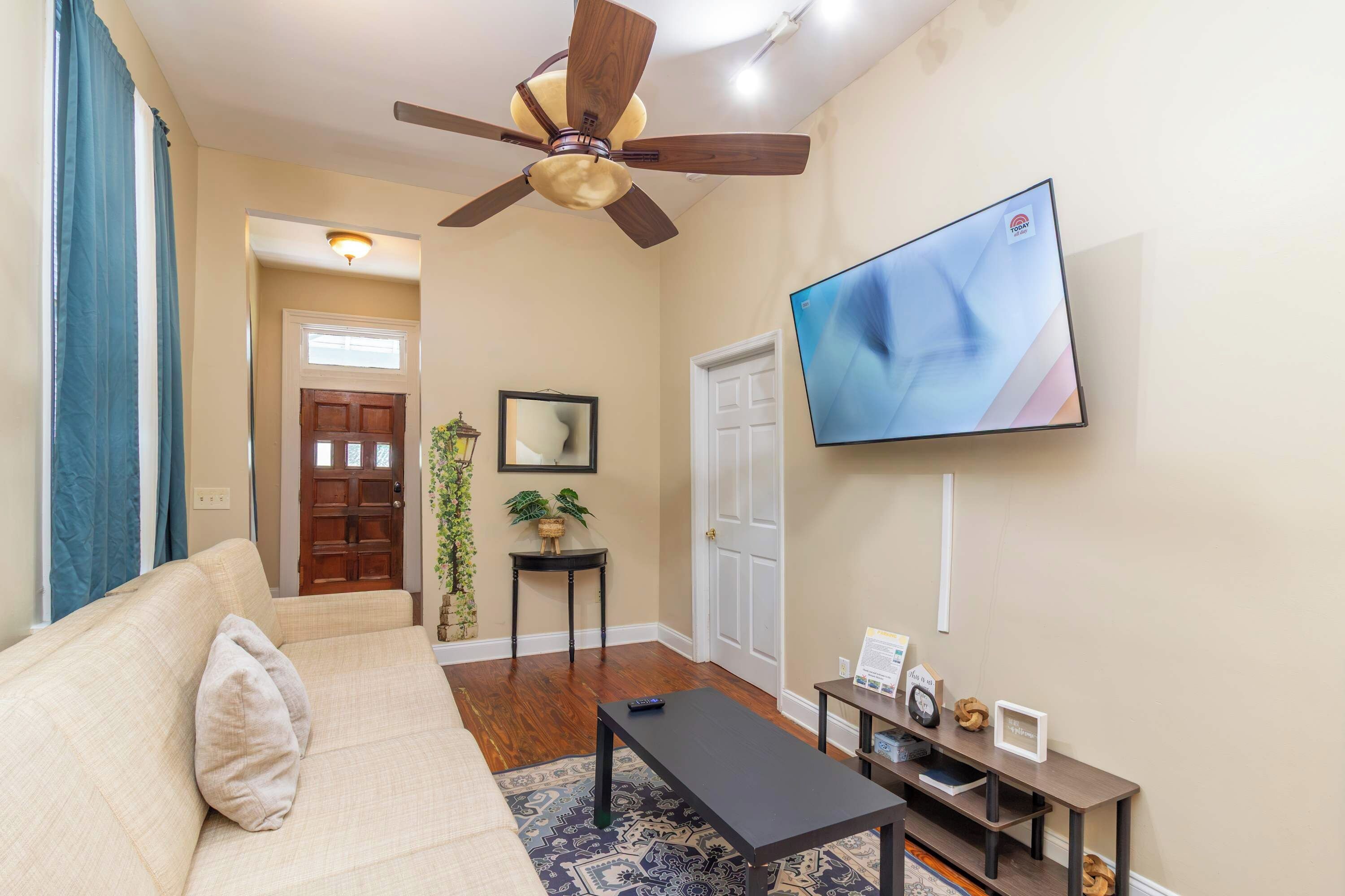 Un salon comprenant un canapé, une table basse, un téléviseur fixé au mur, un ventilateur de plafond et une porte donnant sur une autre pièce.