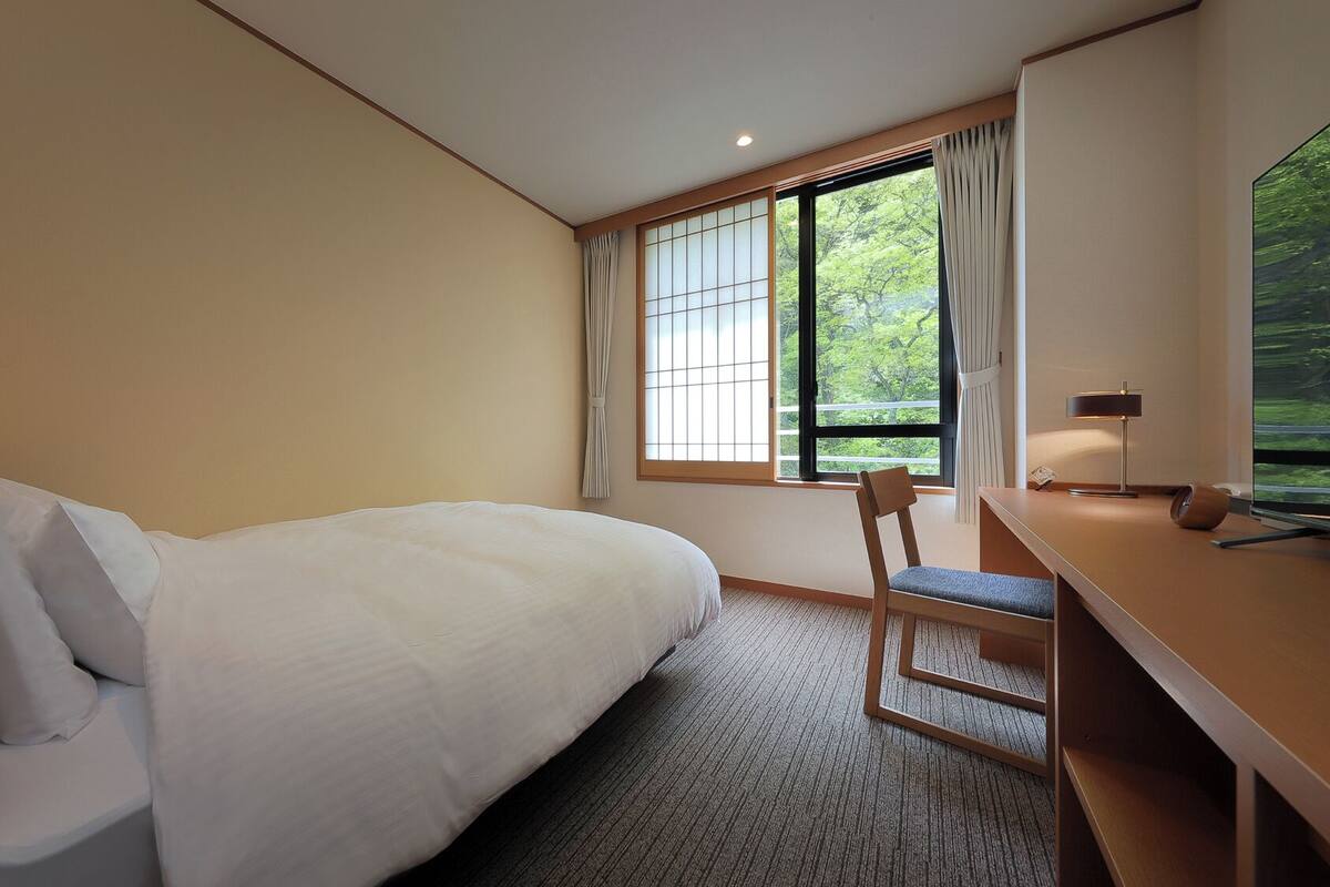 Une chambre d’hôtel avec un lit, un bureau, une chaise et une grande fenêtre donnant sur un paysage verdoyant.
