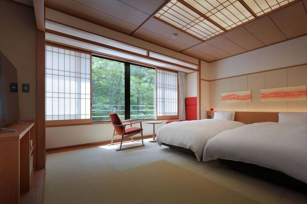 Une chambre d’hôtel moderne avec deux lits, de grandes fenêtres, un fauteuil rouge et des œuvres d’art accrochées au mur.