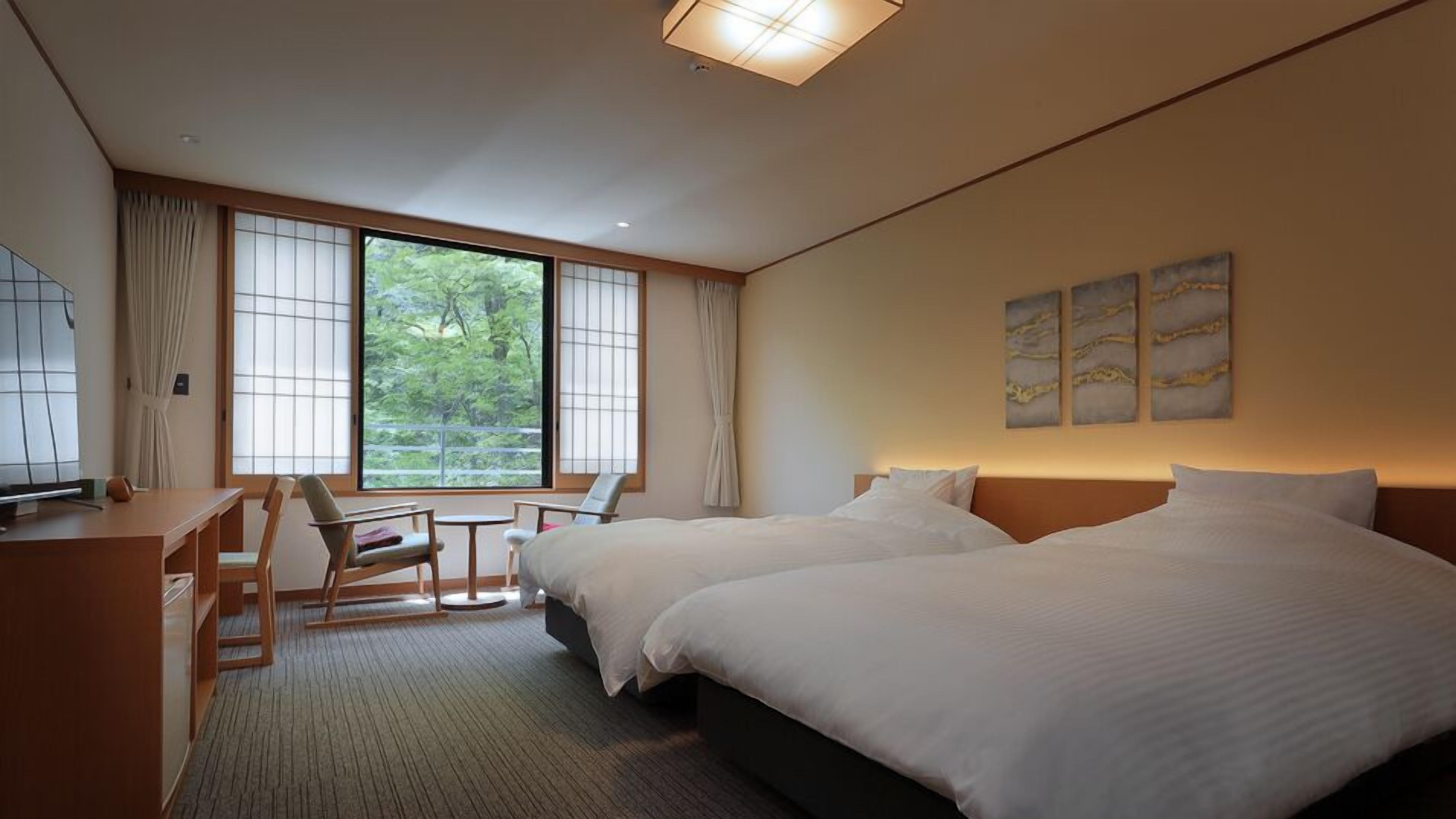 A hotel room with two beds, a desk, a chair, and a view of greenery.