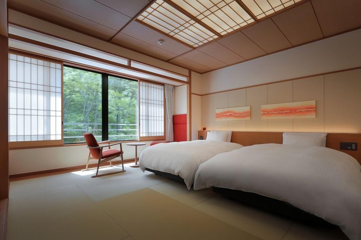 Une chambre d’hôtel moderne avec deux lits, de grandes fenêtres, un fauteuil rouge et des œuvres d’art accrochées au mur.