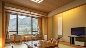 Una habitación tradicional japonesa con suelo de tatami, una mesa baja, sillas, un televisor y vistas a las montañas a través de amplias ventanas.