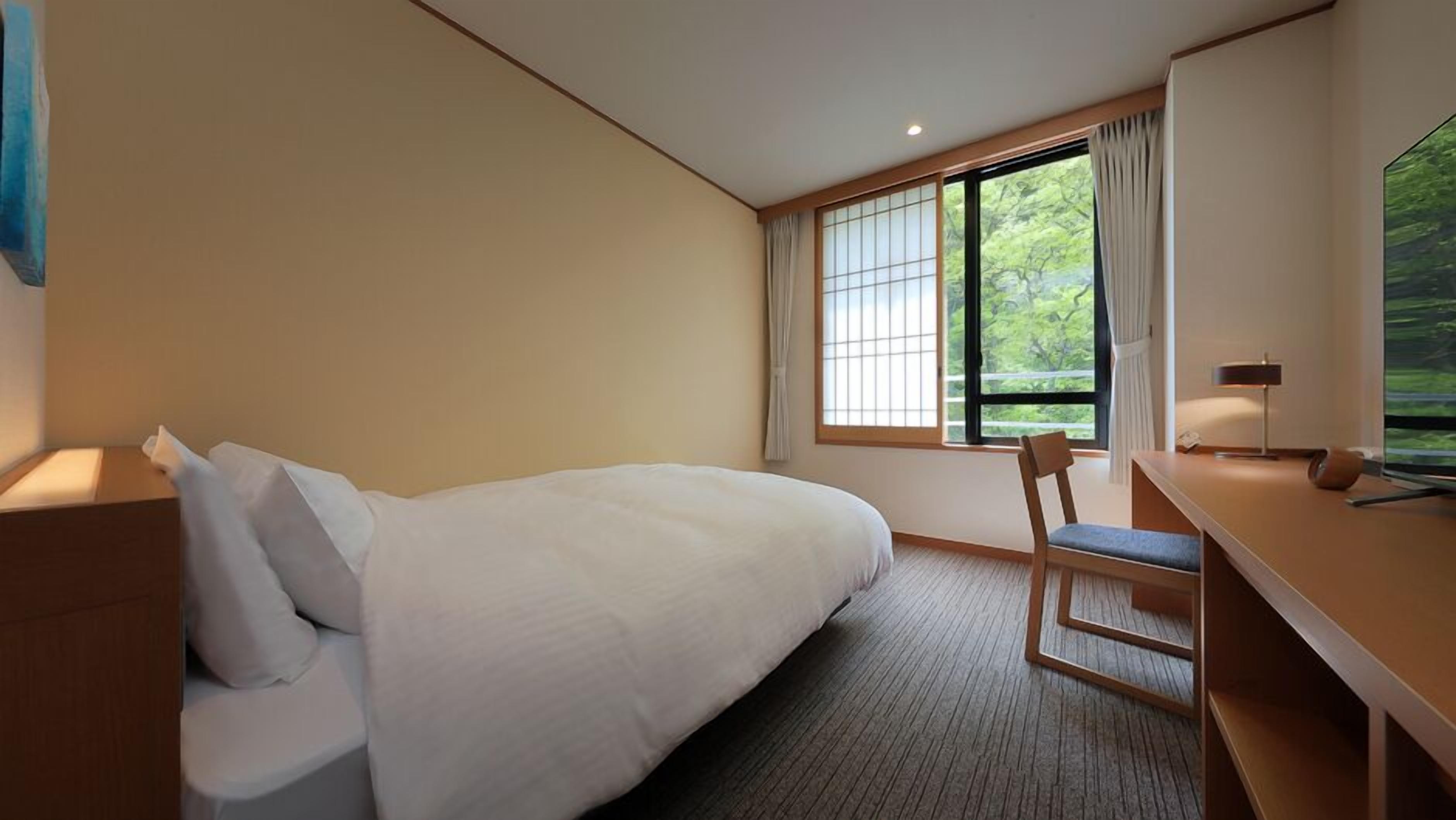 A hotel room with a bed, a desk, a chair, a window with curtains, and a view of greenery.
