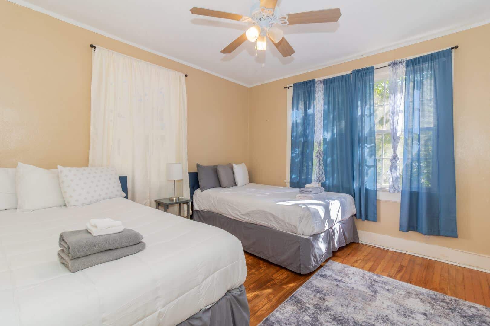 Habitación de hotel con dos camas, ventilador de techo y ventanas amplias con cortinas azules.