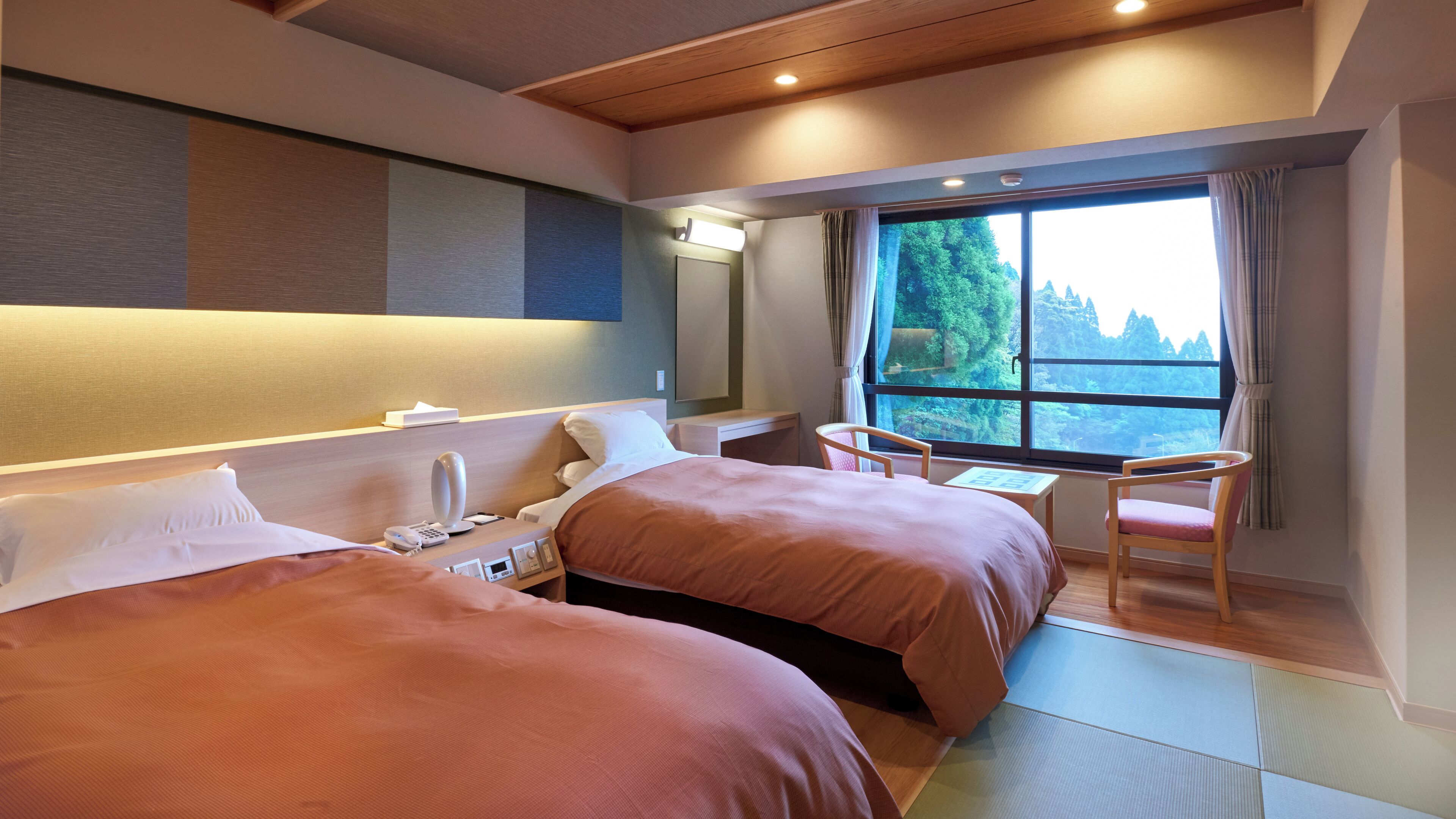 A hotel room with two beds, a desk, a chair, a window with a view of trees, and a ceiling with recessed lighting.
