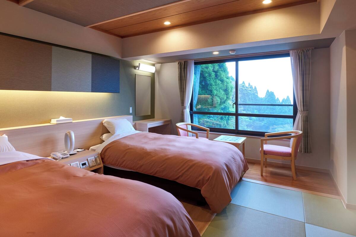 Habitación de hotel con dos camas, un escritorio, una silla, un ventanal con vistas a árboles e iluminación empotrada en el techo.