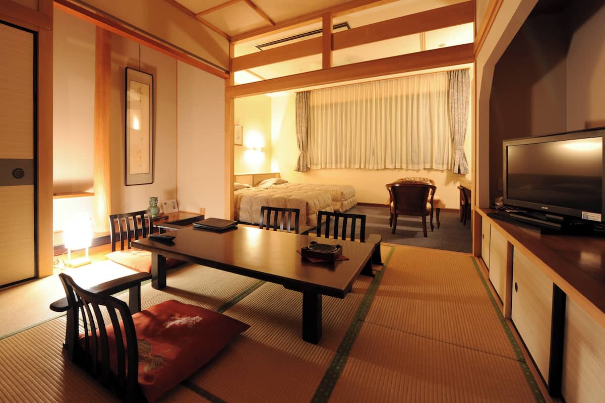 Una habitación tradicional japonesa con suelo de tatami, una mesa baja con sillas, una cama con dosel y un televisor sobre un soporte de madera.