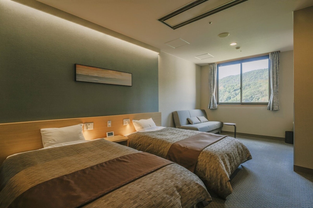 Habitación de hotel con dos camas, una silla, una mesita, ventana con vistas y un cuadro en la pared.