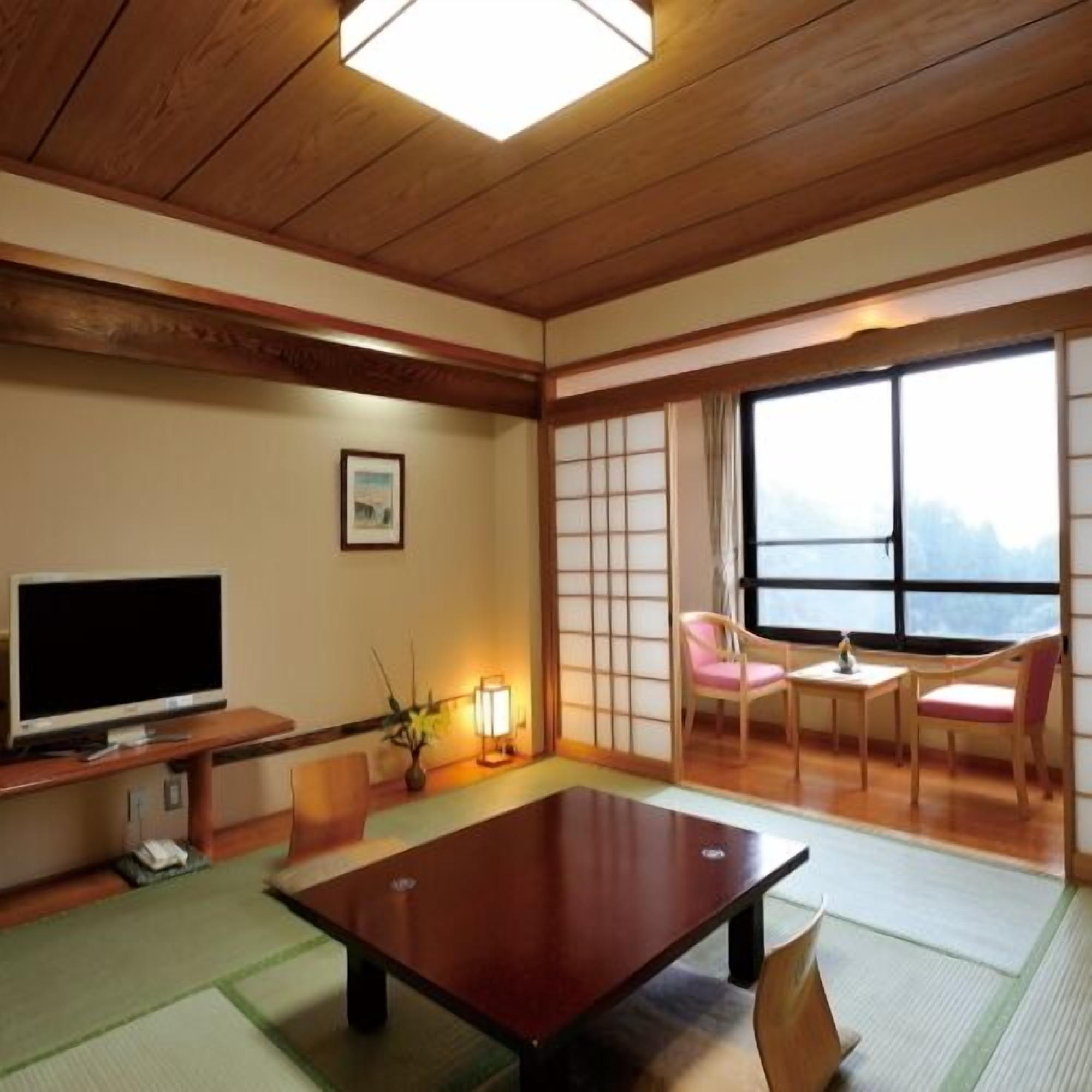 Una habitación japonesa tradicional con suelo de tatami, una mesa baja, sillas, un televisor y un ventanal grande.