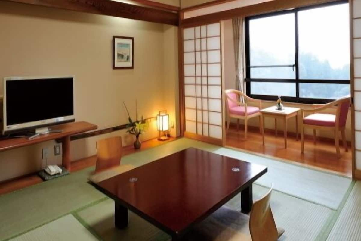 Una habitación japonesa tradicional con suelo de tatami, una mesa baja, sillas, un televisor y un ventanal grande.