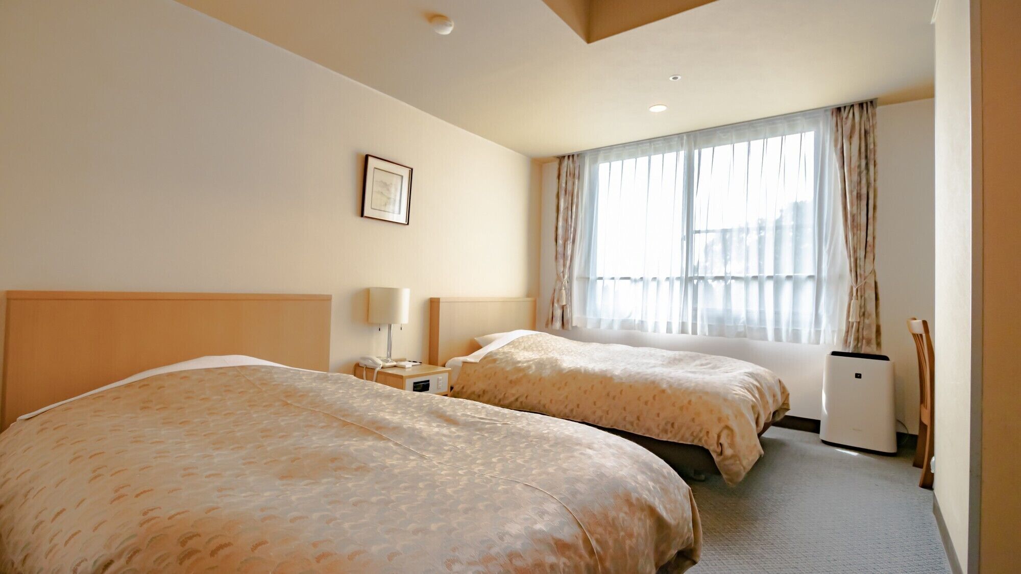 Habitación de hotel con dos camas, una mesita de noche con lámpara, un equipo de aire acondicionado pequeño y una ventana con cortinas.
