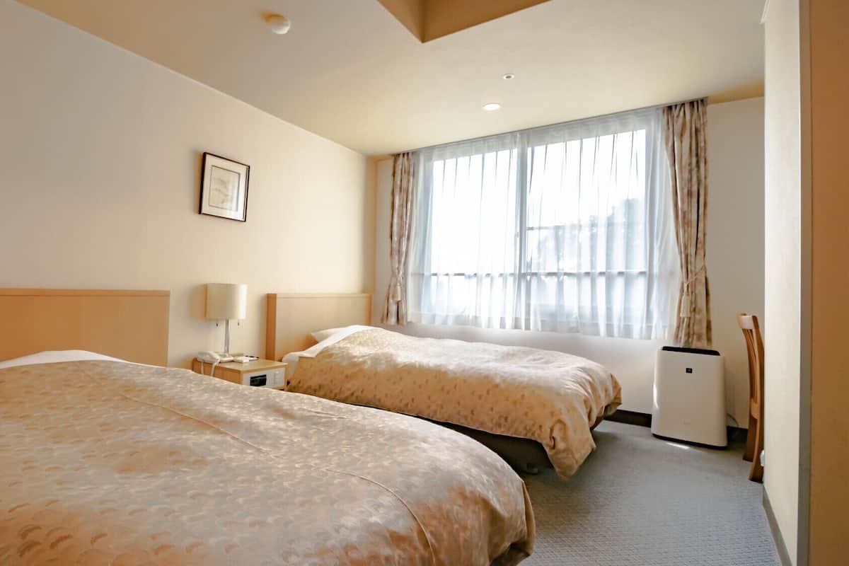 Habitación de hotel con dos camas, una mesita de noche con lámpara, un equipo de aire acondicionado pequeño y una ventana con cortinas.