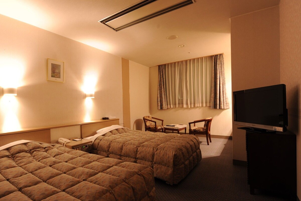 Ein Hotelzimmer mit zwei Betten, einem Schreibtisch, einem Stuhl, einem Fernseher und einem Bild an der Wand.