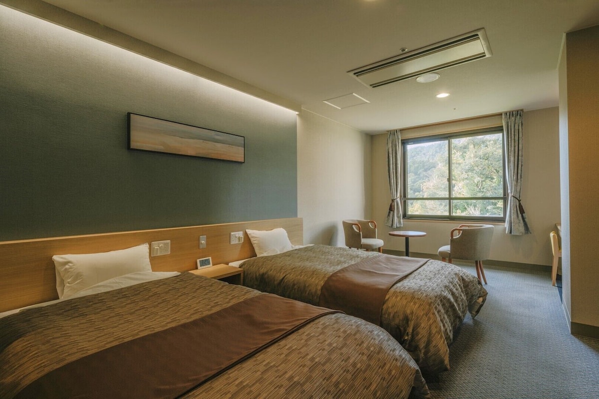 Habitación de hotel con dos camas, una mesita, una silla, un ventanal con cortinas y un cuadro en la pared.
