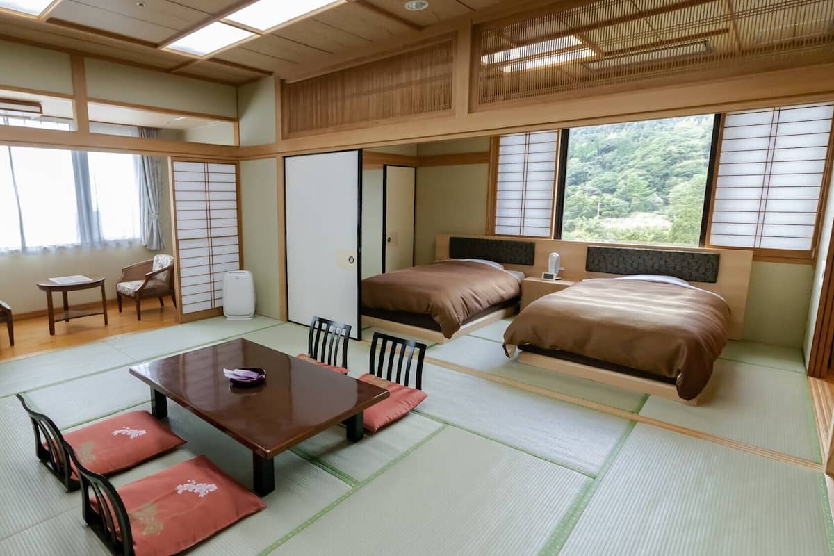 Una habitación japonesa tradicional con suelo de tatami, una mesa baja con cojines, una cama con cabecero y puertas correderas.